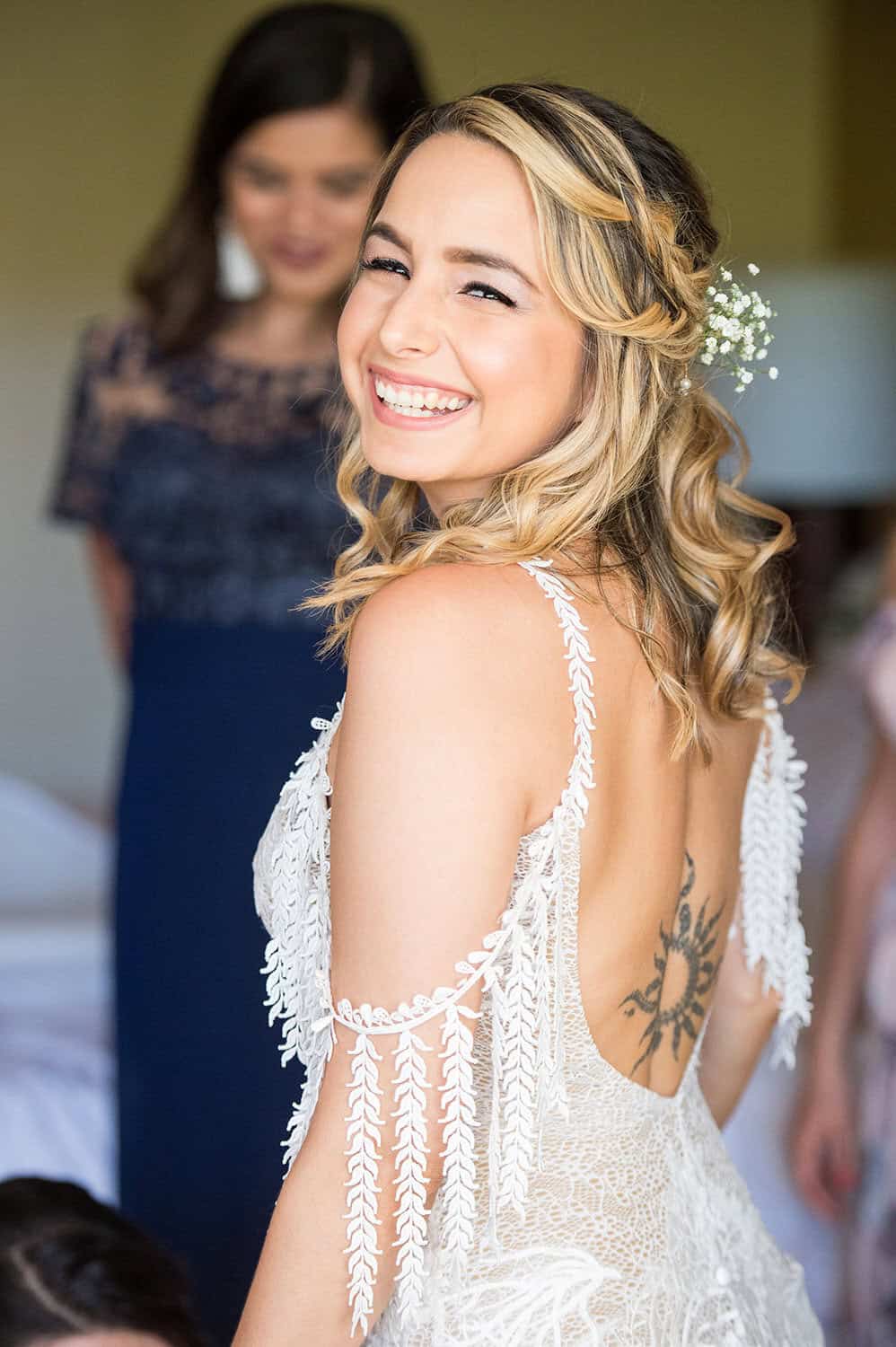 A smiling bride in a white lace dress with an intricate back, featuring a tattoo on her back. Others are in the background, slightly blurred.