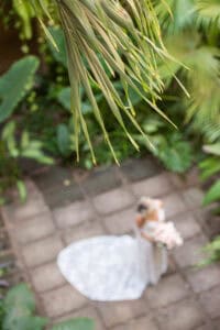 Bride in white gown holding a bouquet, standing on a tiled pathway surrounded by lush greenery, viewed from above through palm leaves.