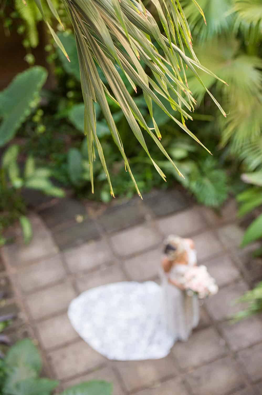 Bride in white gown holding a bouquet, standing on a tiled pathway surrounded by lush greenery, viewed from above through palm leaves.