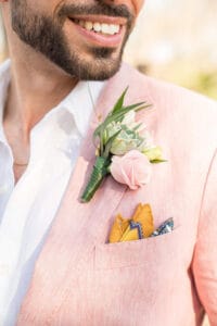 Close-up of a smiling groom wearing a pink suit jacket with a boutonniere of a succulent and pink roses, a white shirt, and patterned pocket squares.
