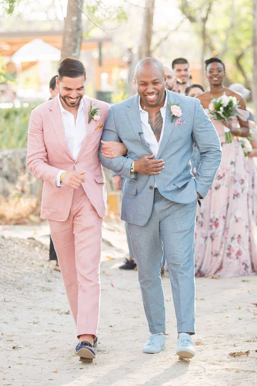 Two men in suits walk arm in arm, smiling and laughing, during an outdoor event. Other people and trees are visible in the background.