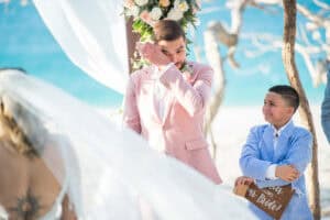 A groom in a pink suit wipes his tears during a beach wedding ceremony. A young boy holding a sign that reads Here comes the bride stands beside him.