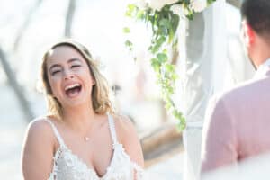 Bride in a white lace dress laughing in an outdoor setting, with a groom in a light-colored suit standing nearby. White flowers and greenery are nearby.