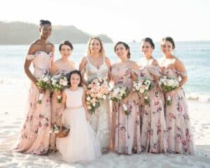 A bride stands with five bridesmaids in pink floral dresses and a flower girl on a sandy beach with the ocean in the background.