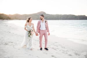 A newlywed couple walks hand in hand on a sandy beach, smiling. The bride wears a white lace wedding dress, and the groom wears a light pink suit.