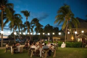 People seated at tables under string lights on a lawn, surrounded by palm trees with a building in the background, during twilight.