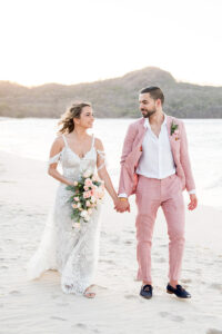 A newlywed couple walks hand in hand on a sandy beach, smiling. The bride wears a white lace wedding dress, and the groom wears a light pink suit.
