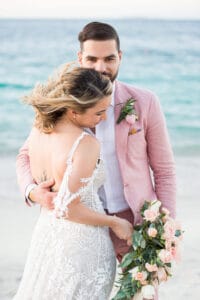 A couple dressed in wedding attire stands on a beach. The woman, in a lace dress, holds a bouquet of roses while the man, in a pink suit, stands behind her.