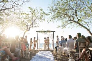Outdoor wedding ceremony on a beach with an ocean backdrop. Two people are getting married under an archway, surrounded by guests on either side of the aisle.