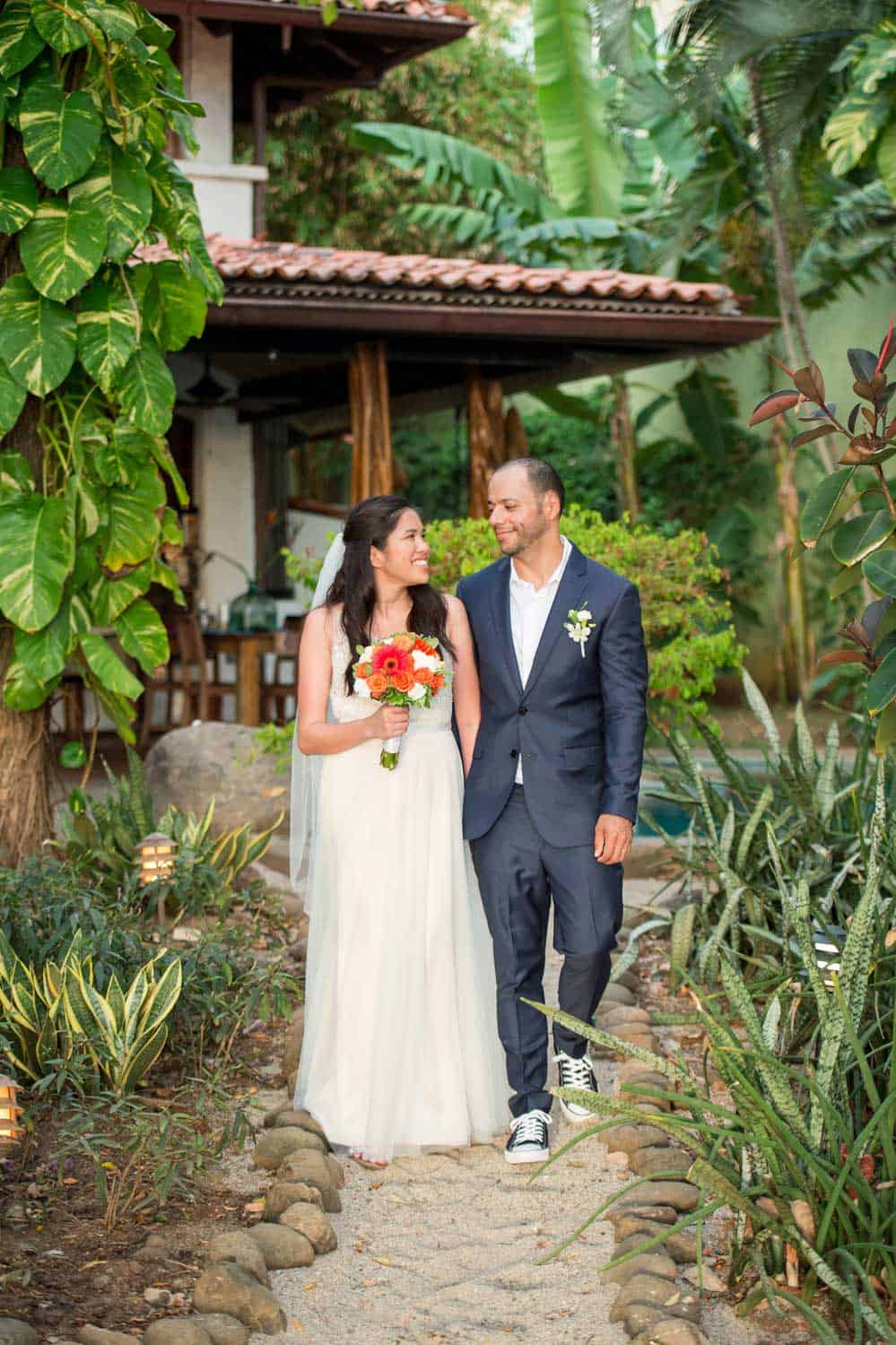 A smiling bride and groom are walking down a garden path. The bride is holding a bouquet of flowers, and the groom is dressed in a suit with sneakers.