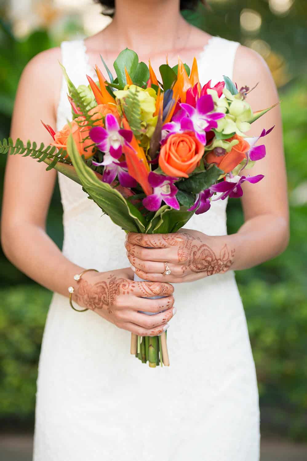 A person in a white dress holds a vibrant bouquet of mixed flowers with detailed henna designs on their hands.