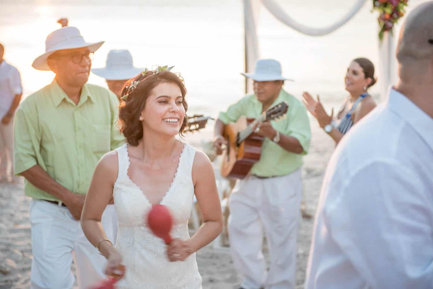 A bride in a white dress smiles while playing maracas, accompanied by musicians and guests, at an outdoor beach wedding.