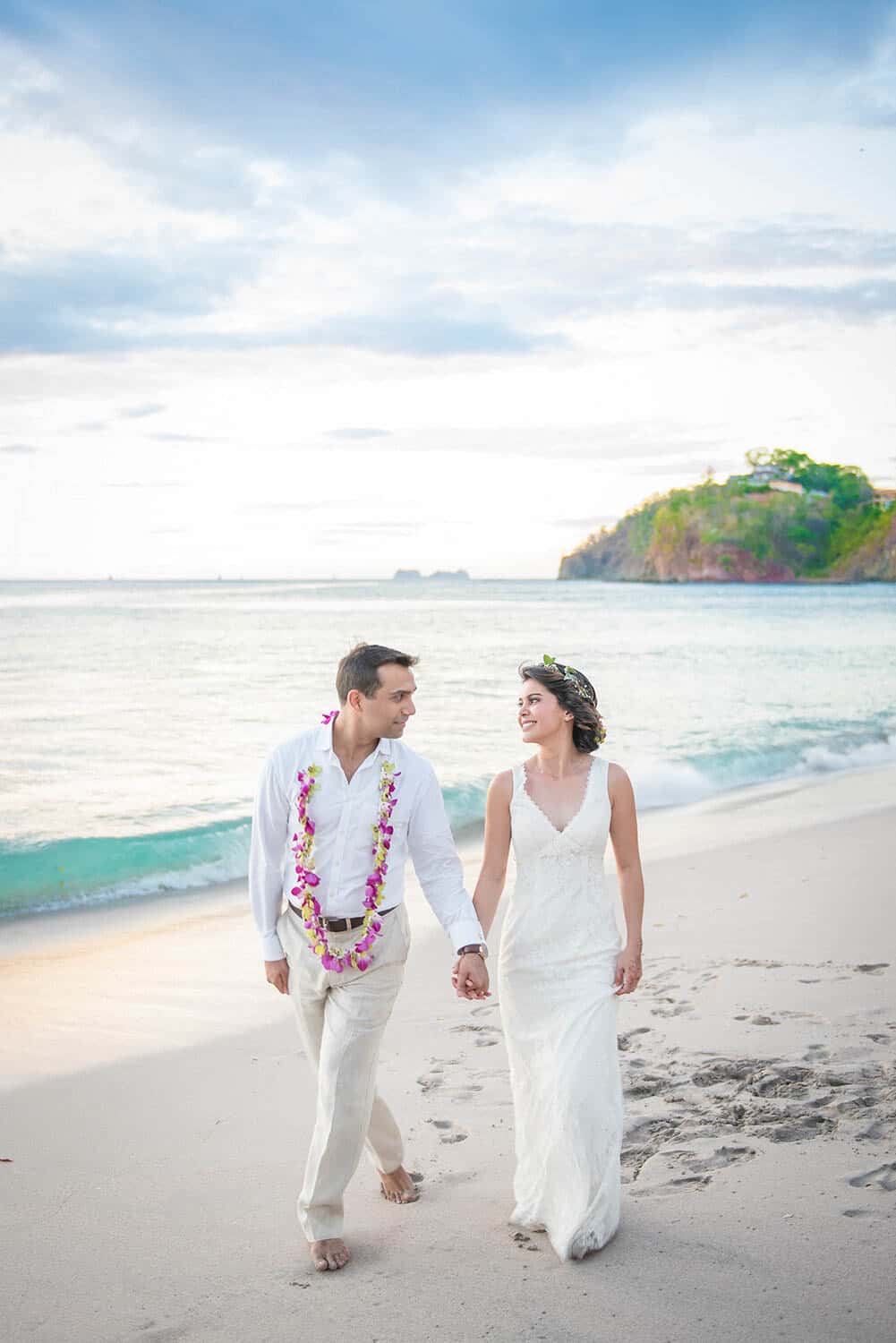 A man wears a white outfit with a flower lei, and a woman wears a white dress and floral headpiece, walking hand-in-hand along a beach.