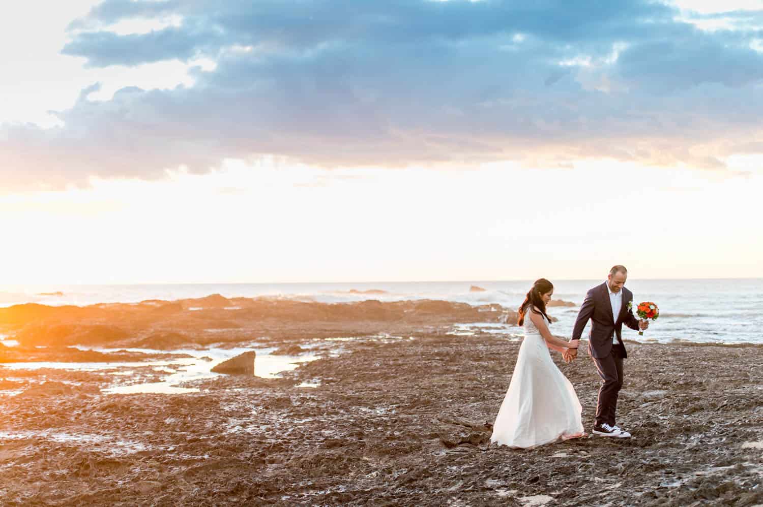 A bride and groom walk hand-in-hand along a rocky shoreline at sunset, with the groom holding a bouquet of flowers.