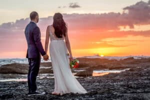 A couple, dressed in wedding attire, stands hand in hand on a rocky shore, watching a vibrant sunset over the ocean. The bride holds a bouquet of flowers.
