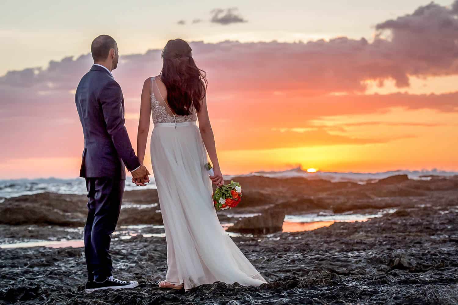 A couple, dressed in wedding attire, stands hand in hand on a rocky shore, watching a vibrant sunset over the ocean. The bride holds a bouquet of flowers.