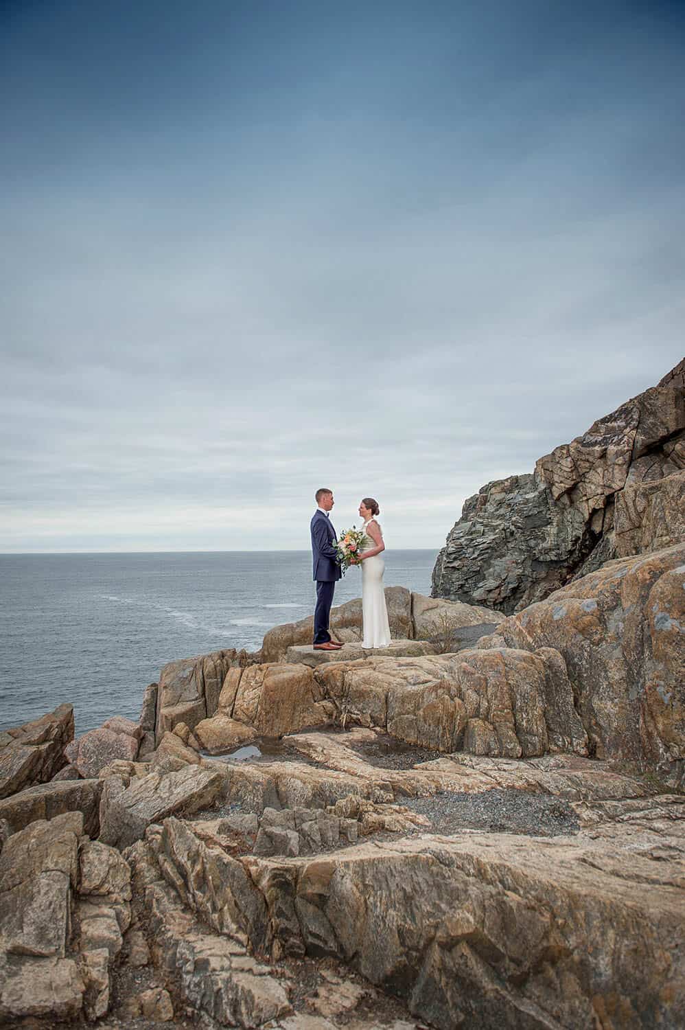 A couple stands on rocky cliffs near the ocean, holding hands and facing each other, with a cloudy sky above.