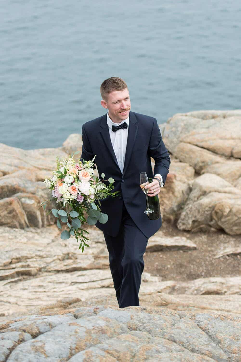 A man in a tuxedo holding a bouquet and a wine bottle walks on rocky terrain by the sea.