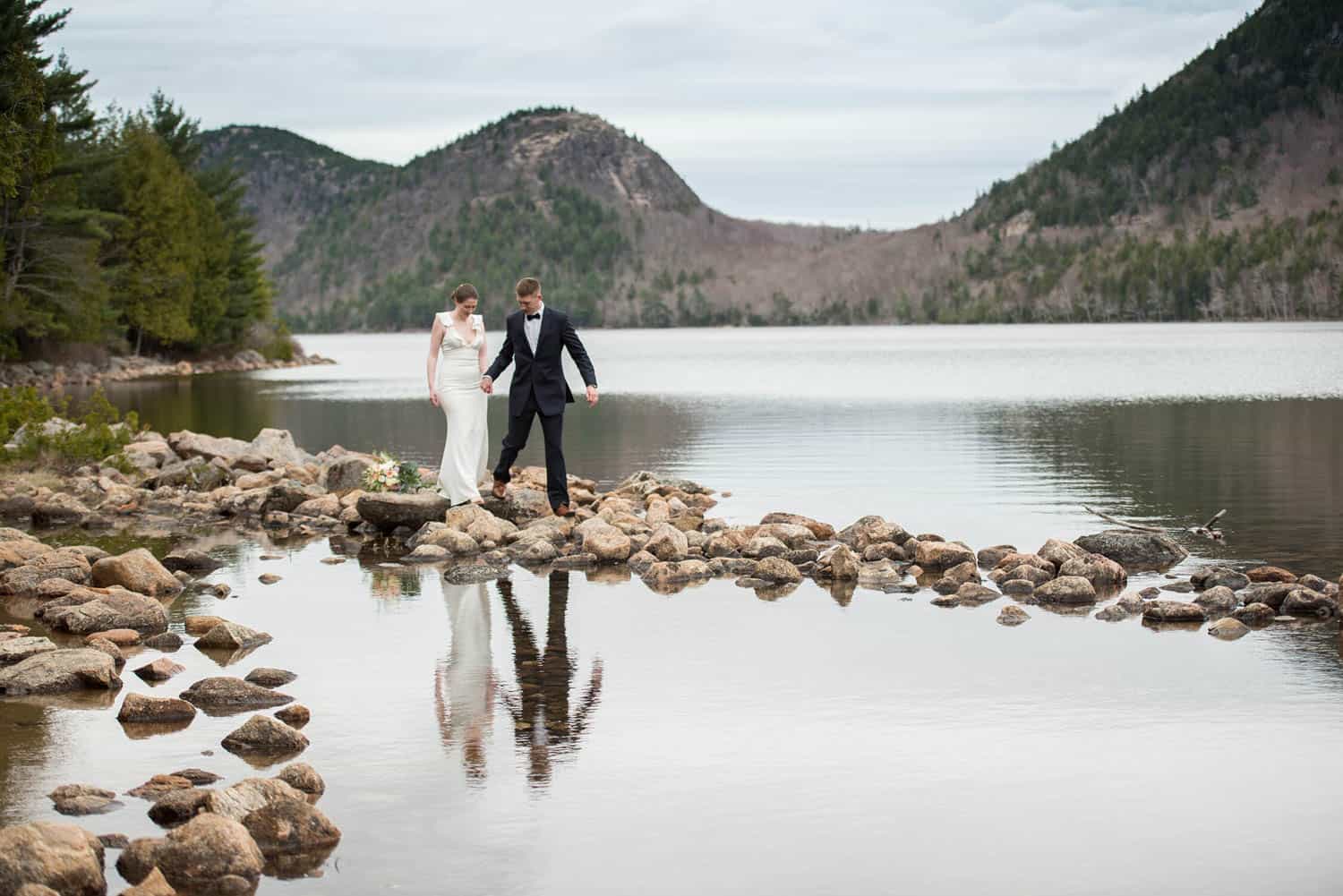 A couple in formal attire walks hand-in-hand by the edge of a serene lake surrounded by rocky shores and forested hills.