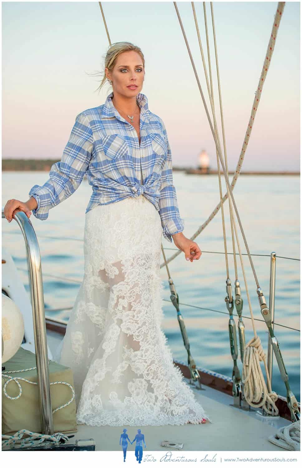 A woman wearing a plaid shirt over a wedding dress stands on a sailboat holding onto a railing with one hand, with calm waters and a distant lighthouse nearby.