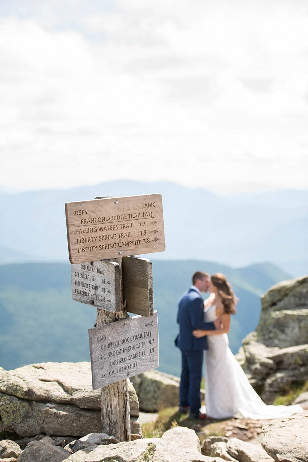 A couple in wedding attire stands closely together and kisses on a rocky mountaintop near a wooden trail sign under a partly cloudy sky.