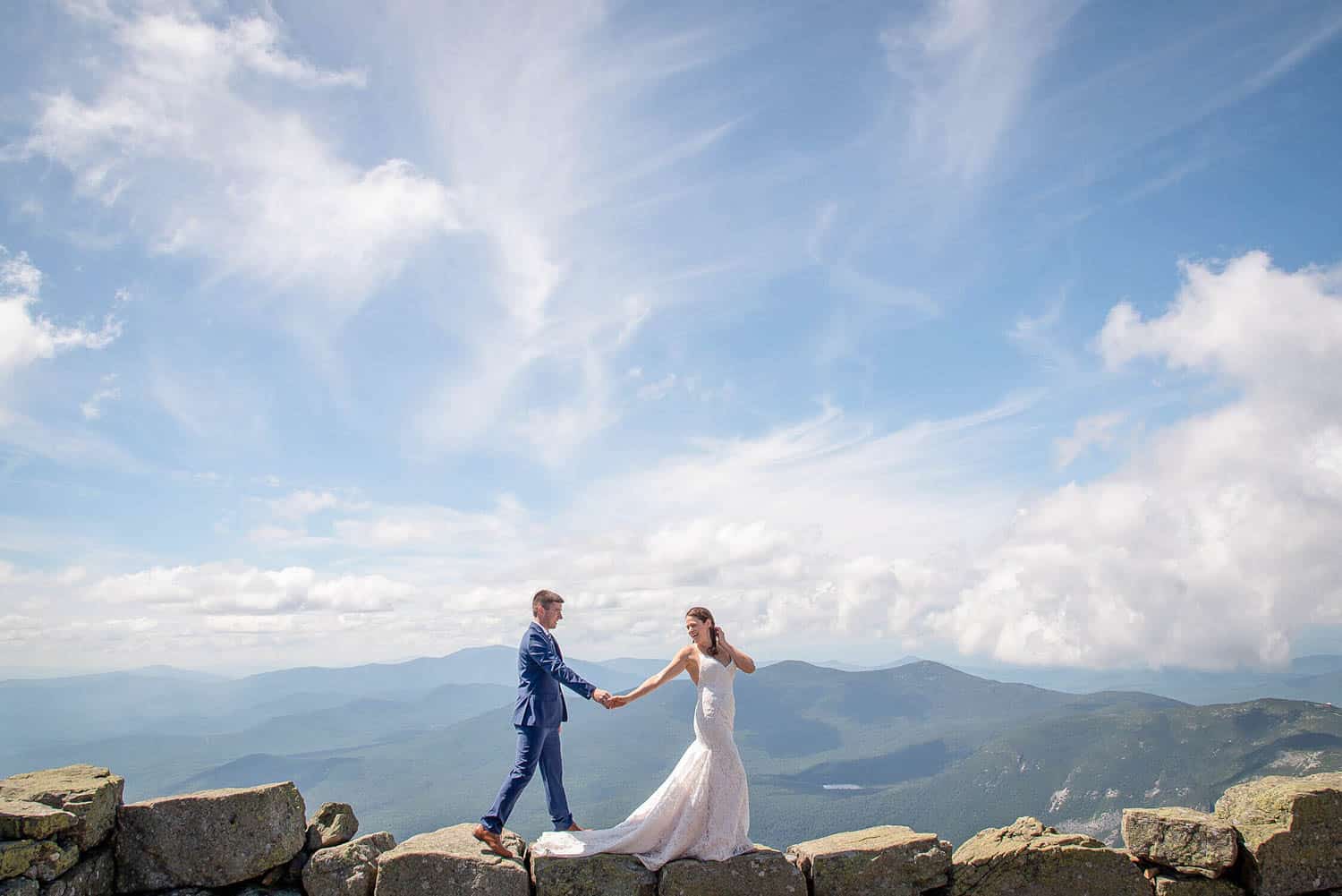 A bride and groom in wedding attire hold hands and walk on a rocky ledge with a vast mountain landscape and a blue sky with clouds in the background.