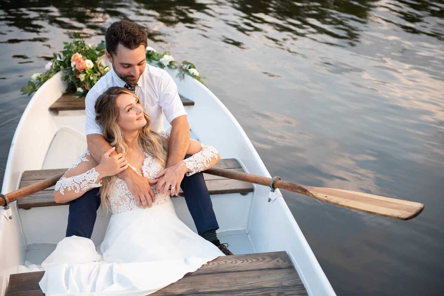 A couple sits in a small white rowboat on a body of water. The woman, in a white lace dress, leans back against the man, who is wearing a white shirt.