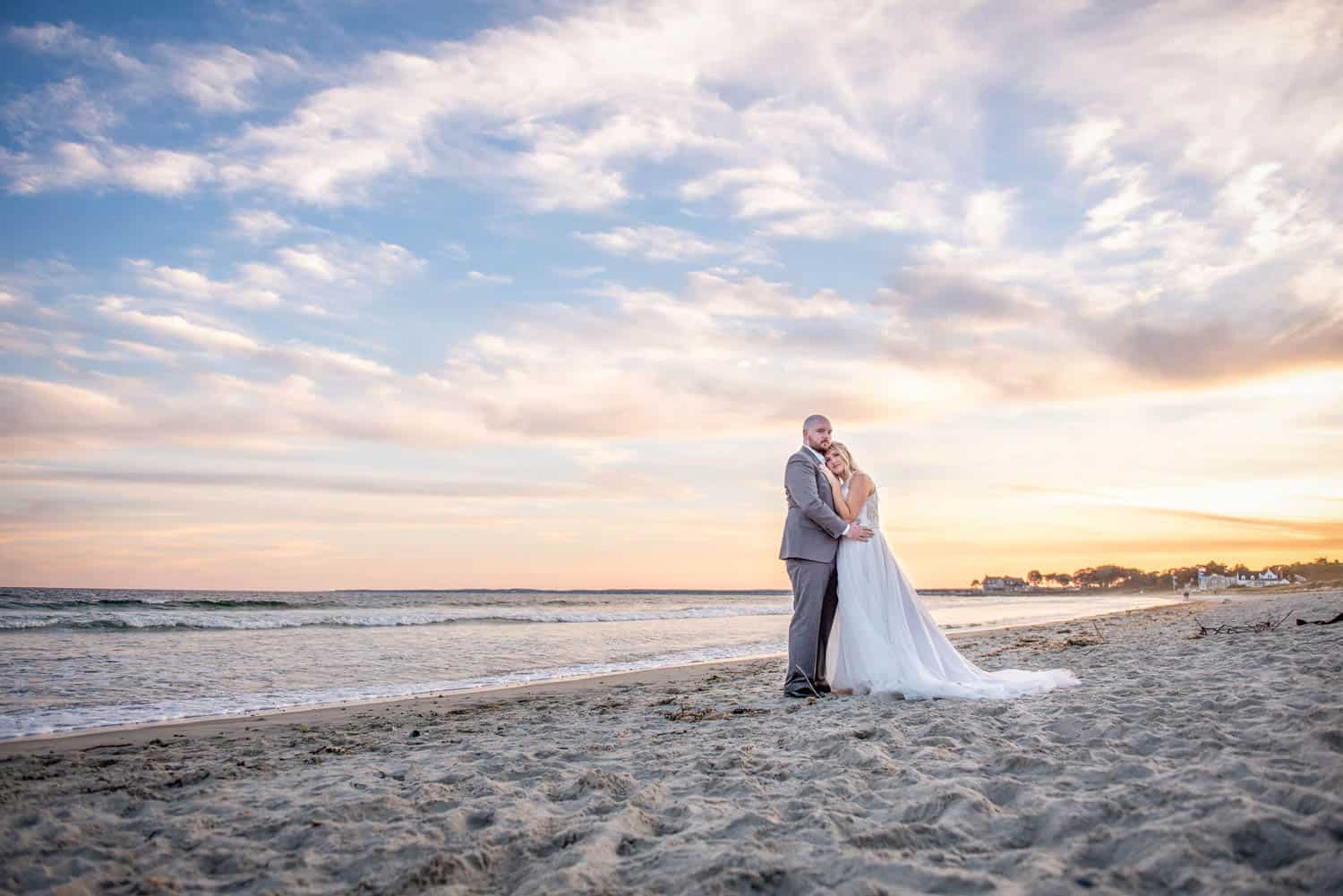 A couple dressed in wedding attire stands on a sandy beach at sunset, with the ocean and a colorful sky in the background.