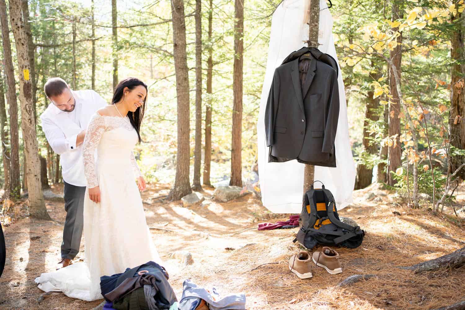 A bride and groom get dressed in a forest. The bride is adjusting her dress, while the groom is putting on his shirt. A suit and shoes hang on a tree nearby.