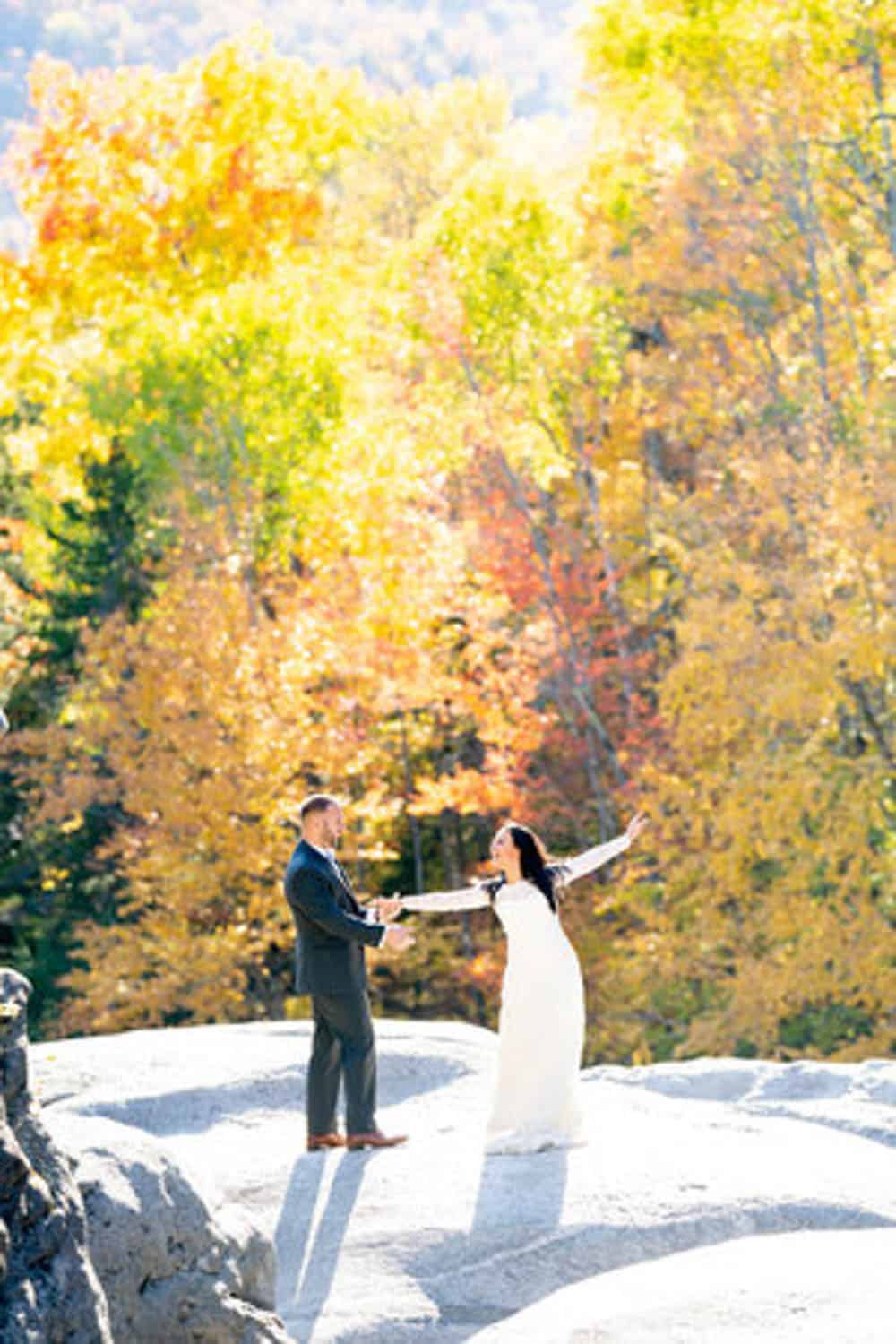 A couple stands on a rocky surface surrounded by autumn trees, with the bride in a white dress extending her arms and the man holding her one hand.