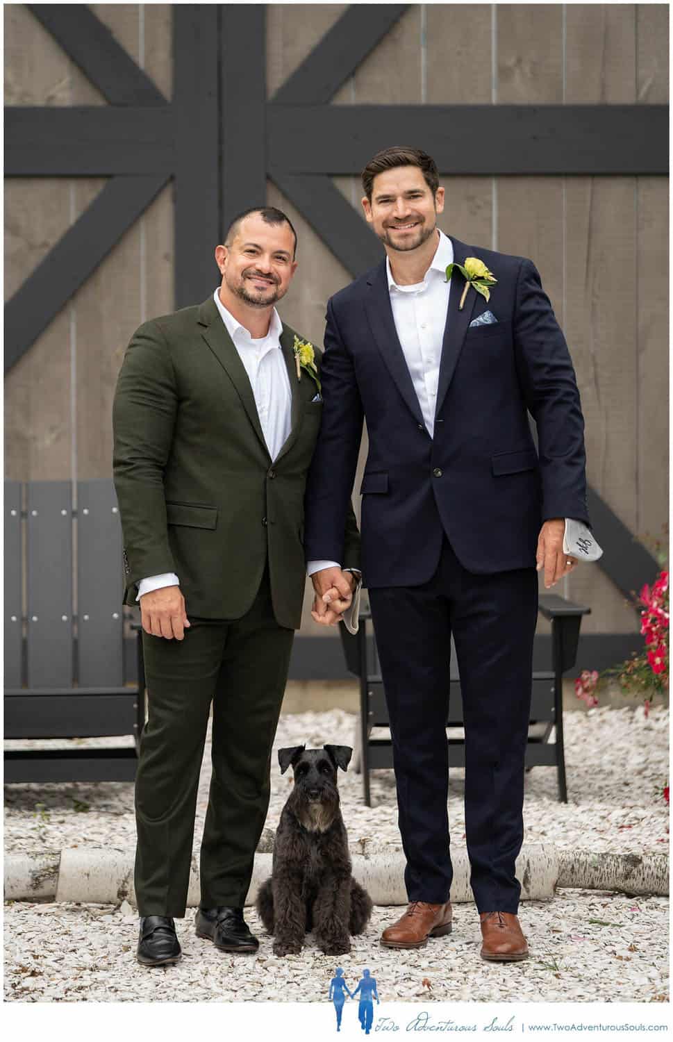 Two men in suits holding hands and smiling, standing with a small black dog in front of them. The background includes a wood fence and outdoor seating.