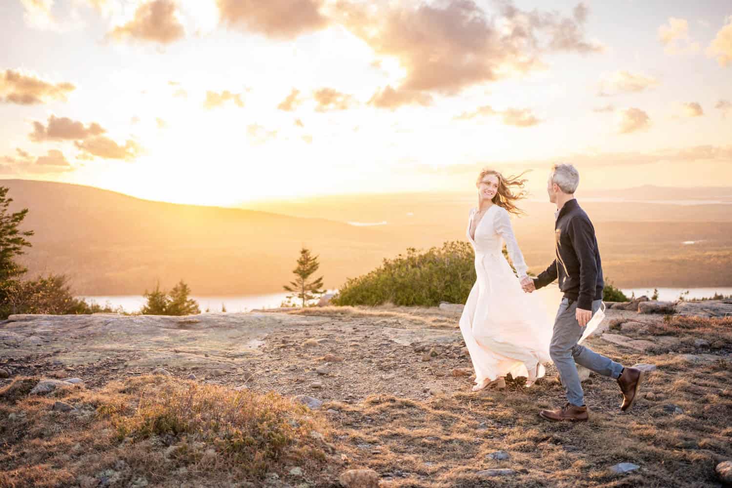 A couple holding hands and smiling while walking on a trail during sunset, with a scenic view of hills and water in the background.