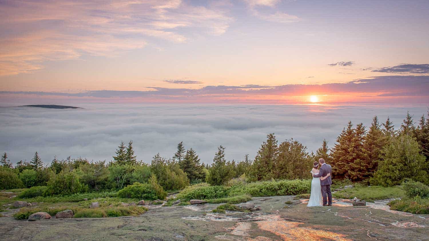 A couple stands on a rocky terrain surrounded by greenery, overlooking a sea of clouds at sunset, with the sun partially visible on the horizon.