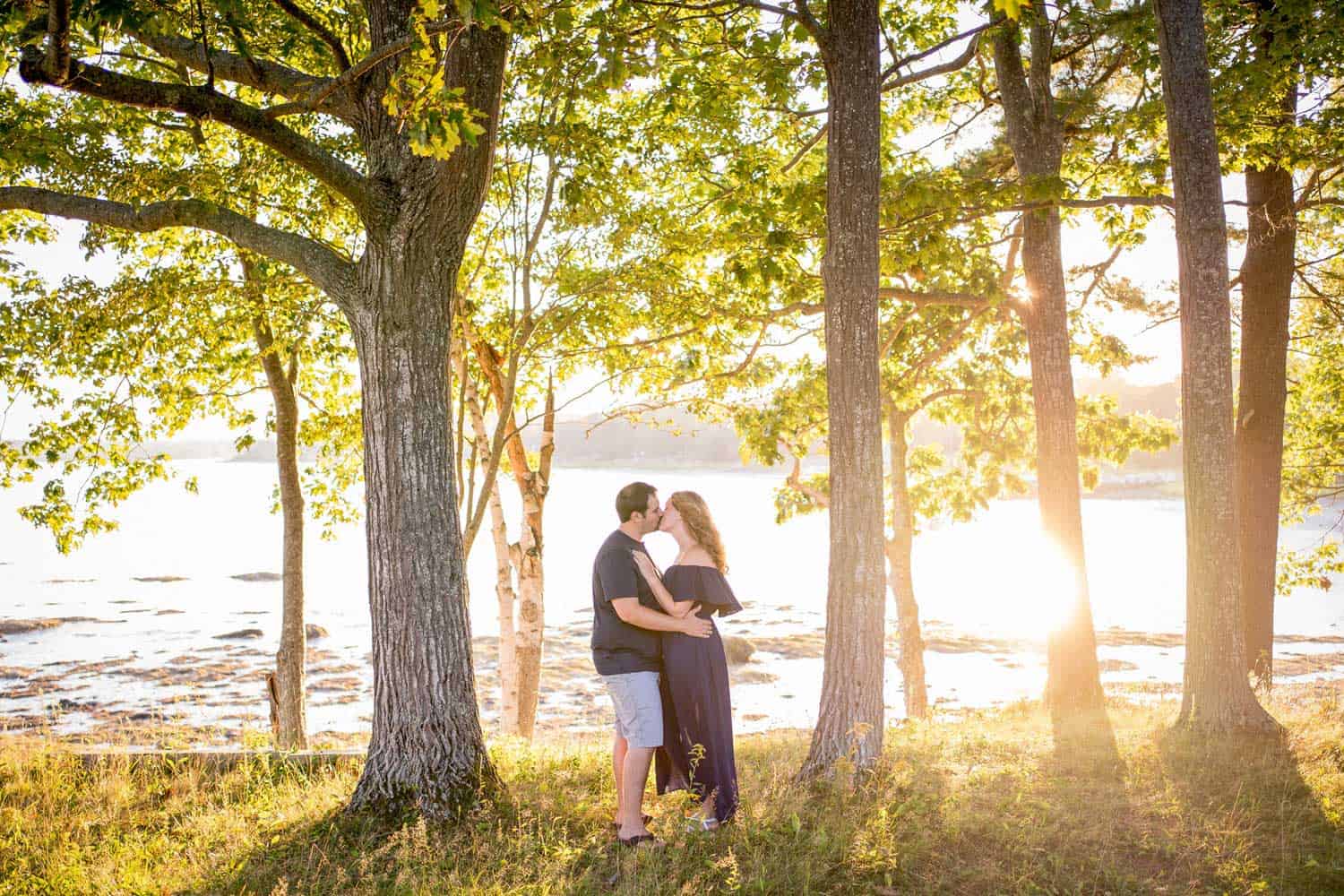 A couple embraces among trees near a body of water at sunset, with sunlight streaming through the branches.