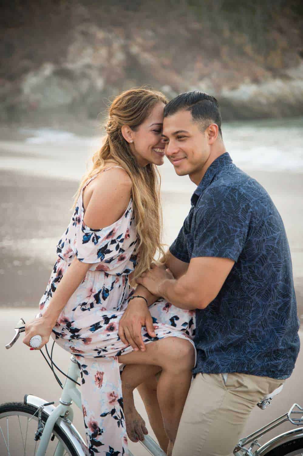 A woman and man smile while sitting on a bicycle on a beach. The woman wears a floral dress, and the man is in a dark shirt and beige pants.