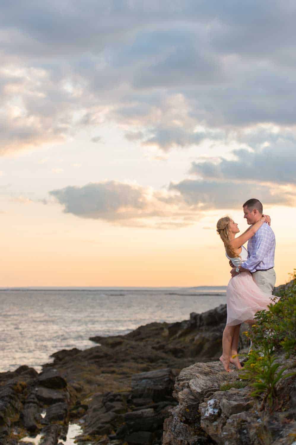 A couple stands on rocky shore, the man holding the woman in his arms as they gaze at each other against a sunset sky by the sea.