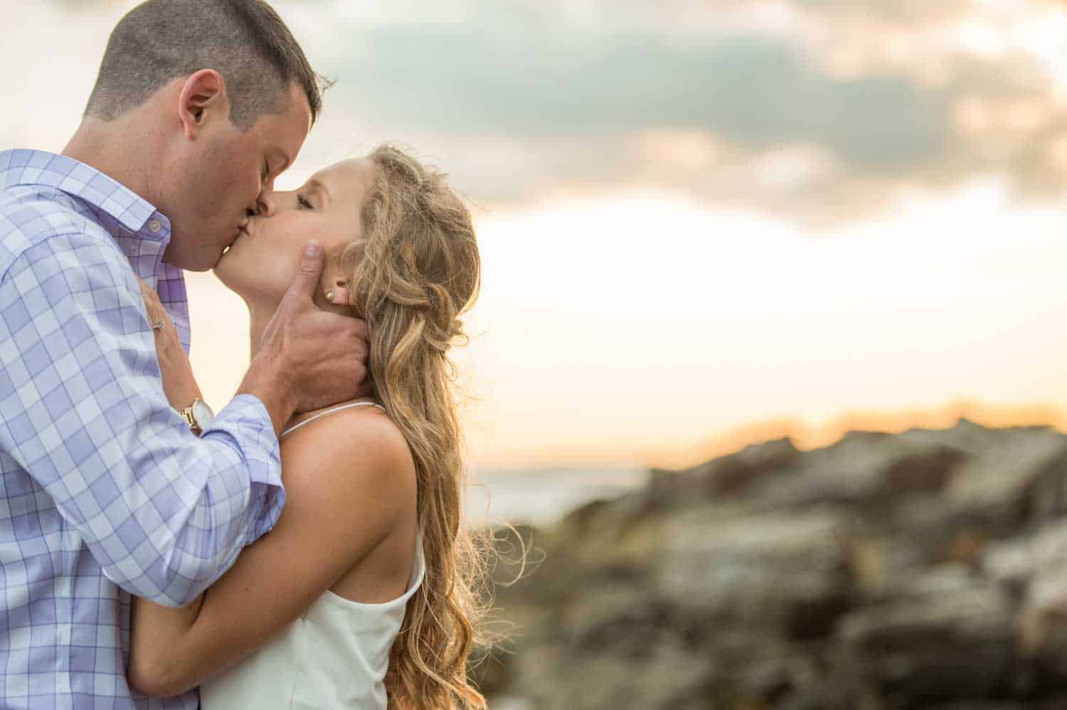 A couple kisses passionately at sunset near a rocky shoreline.