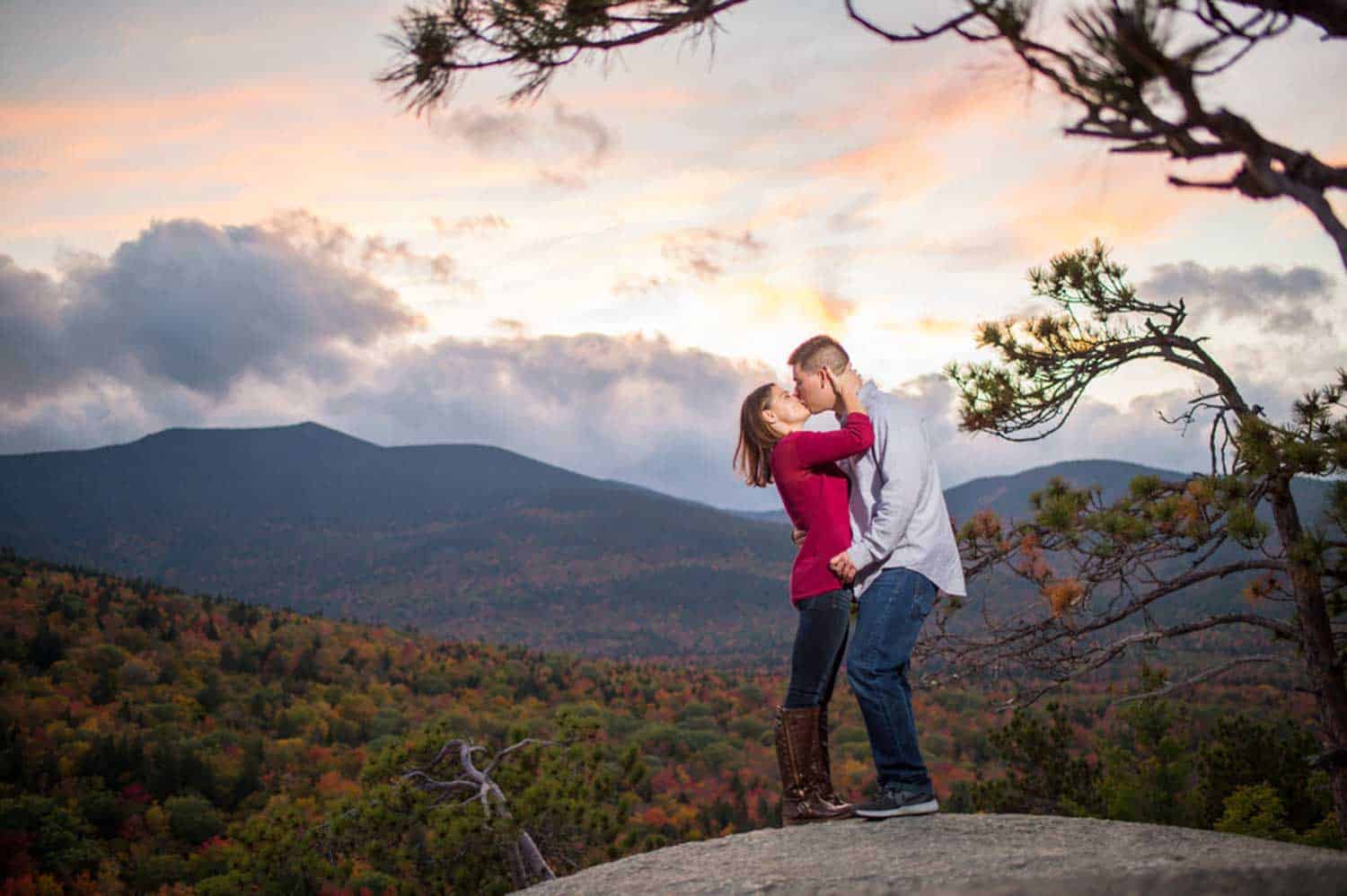 A couple stands on a rocky ledge in front of a mountainous landscape with autumn foliage, sharing a kiss under a colorful evening sky.