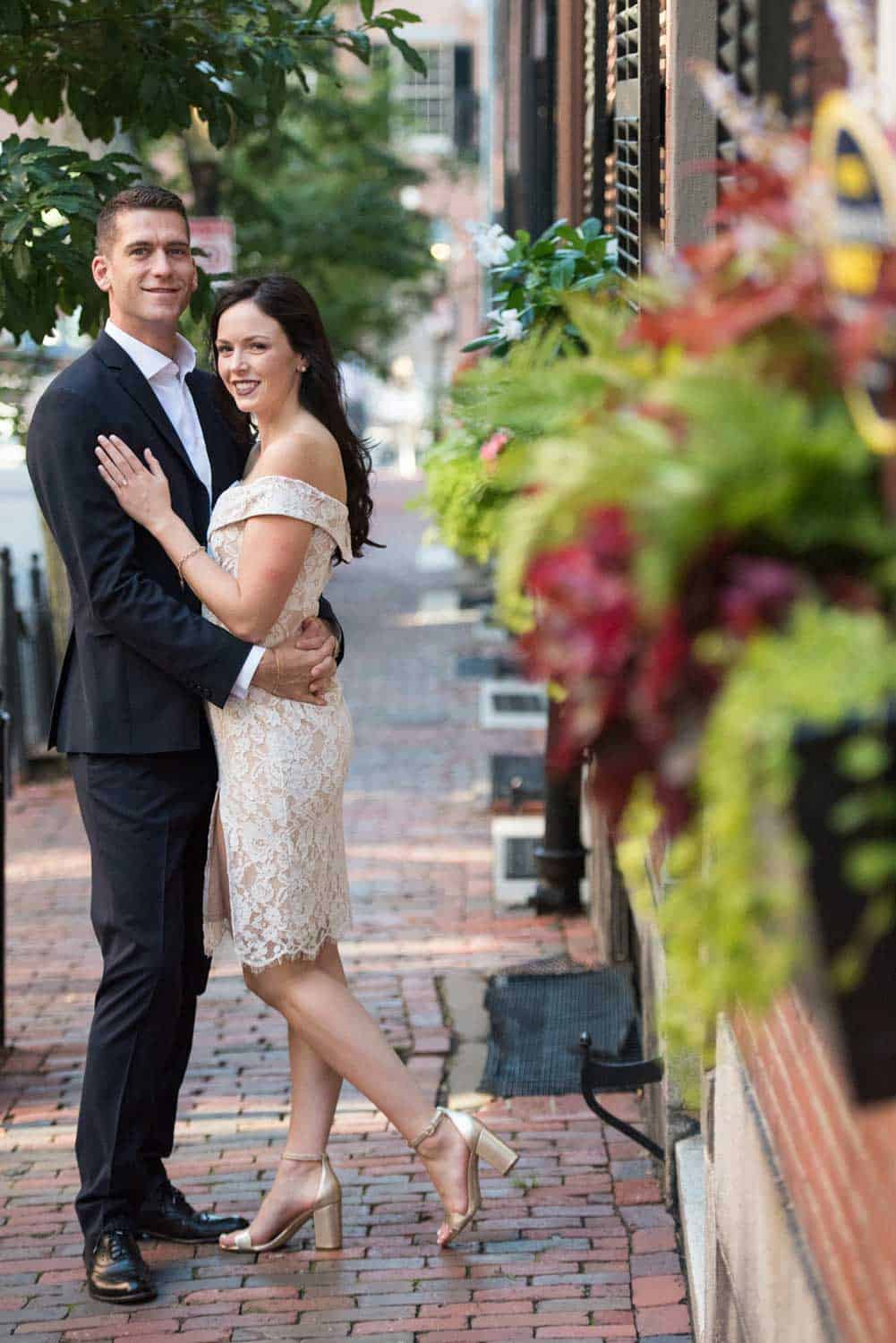 A couple dressed formally, with the man in a suit and the woman in a lace dress, pose together on a brick-paved sidewalk bordered by potted plants and greenery.