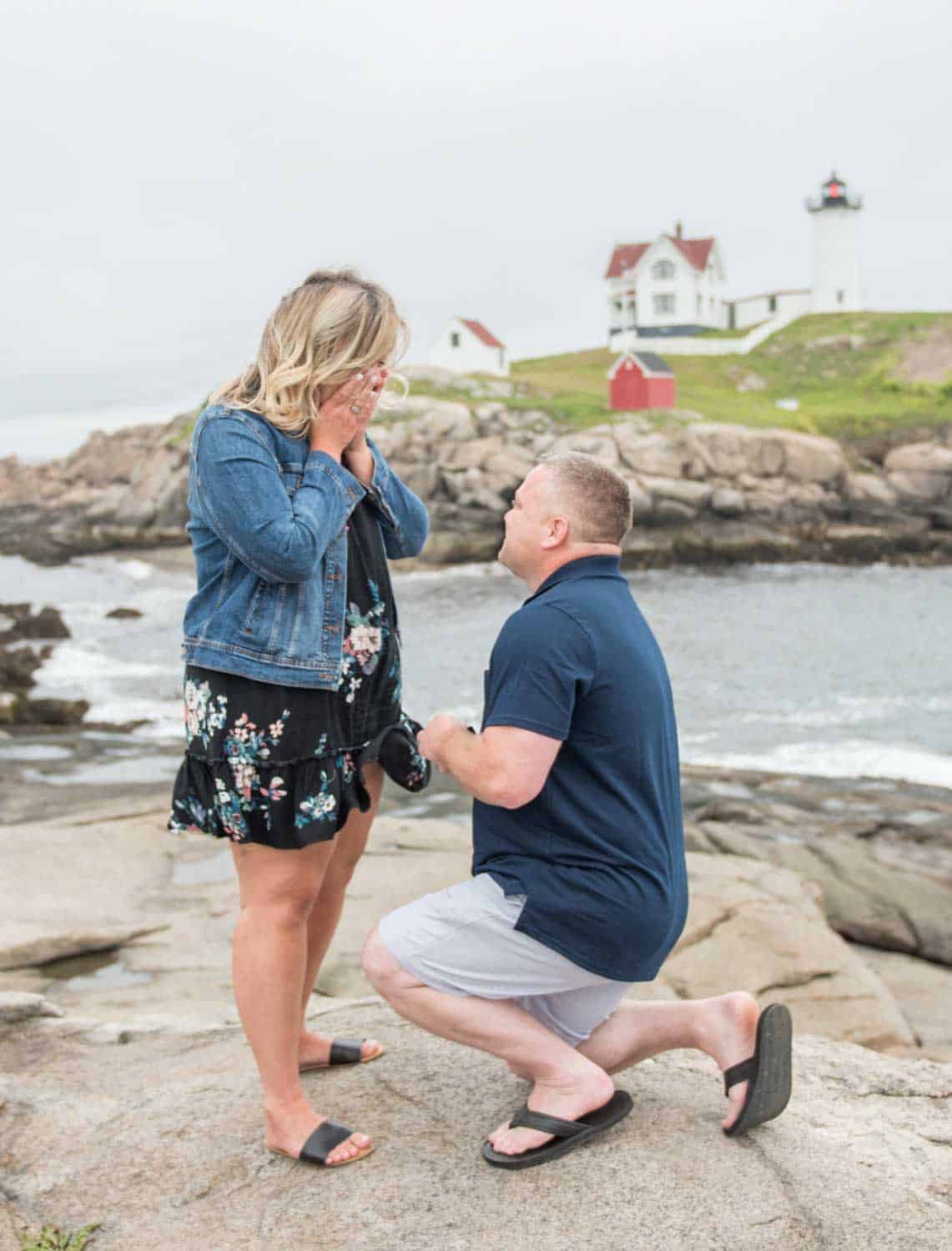 A man kneels on one knee holding a ring, proposing to a surprised woman by the ocean. A lighthouse and house sit atop a rocky hill in the background.