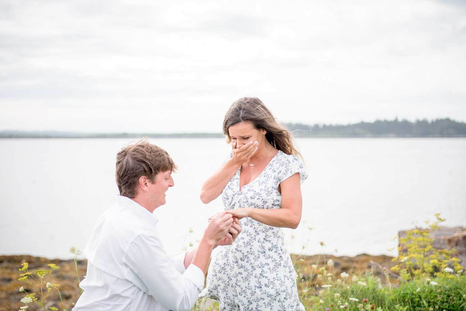 A man kneels and proposes to a surprised woman beside a lake, with greenery in the background. She covers her mouth with one hand and looks at the ring.