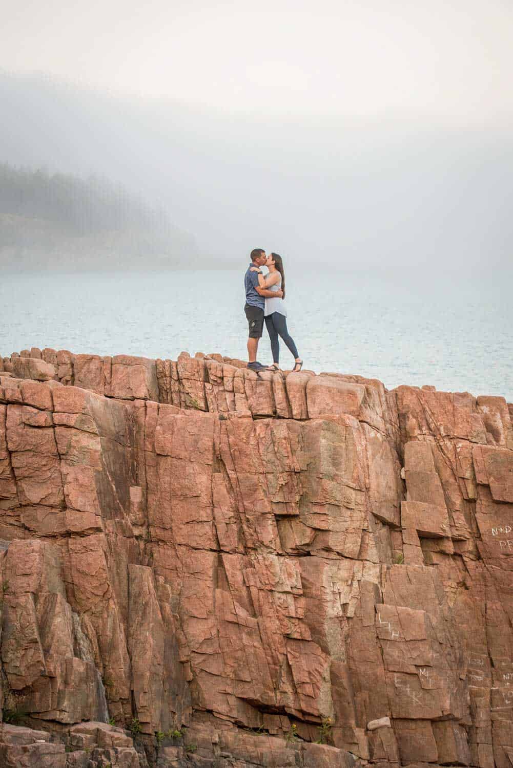 A couple stands on a rocky cliff, embracing and kissing, with a foggy body of water and distant treeline in the background.