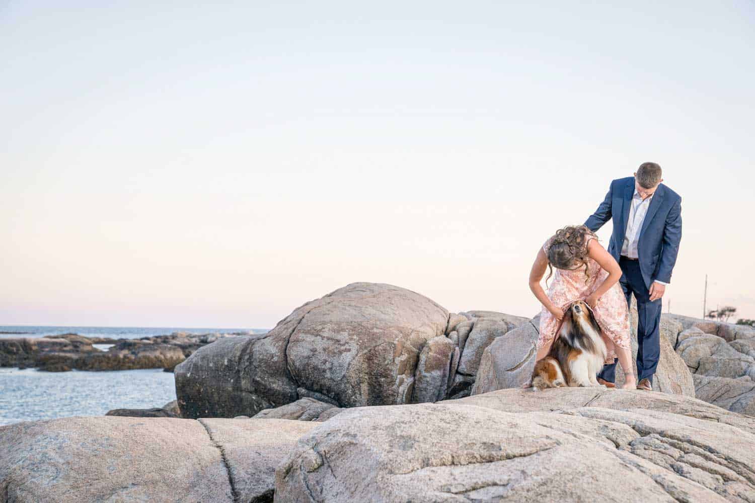 A couple in formal attire stands atop rocky terrain near the sea. A Collie peeks his head out from under the woman's dress.