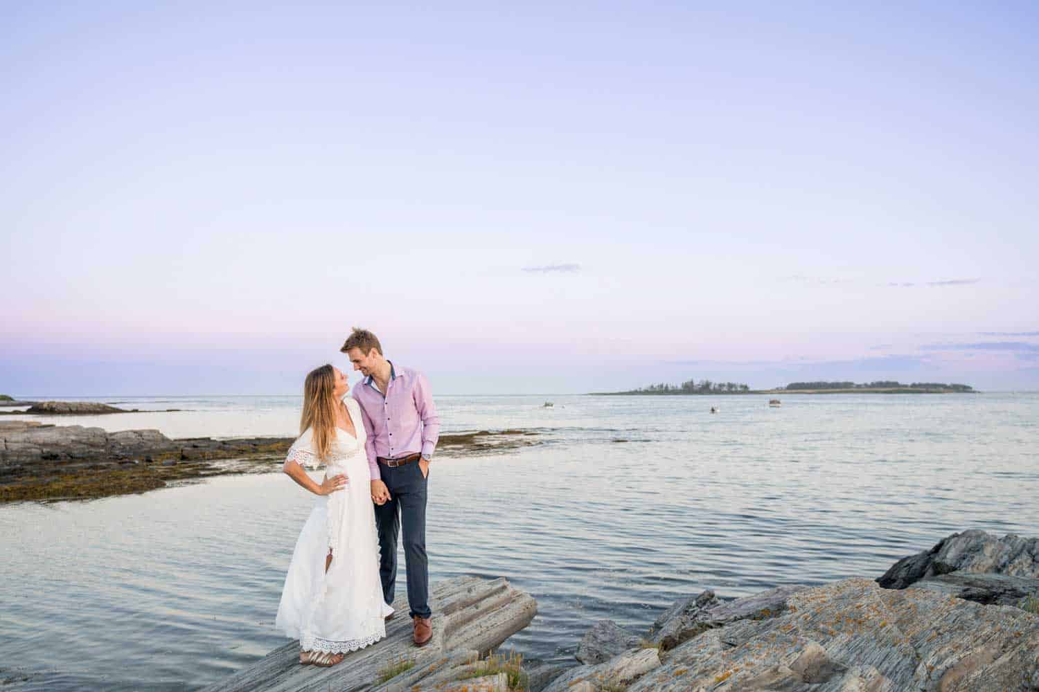 A couple stands on rocks near the shoreline, gazing at each other with a serene ocean and pastel sky in the background.