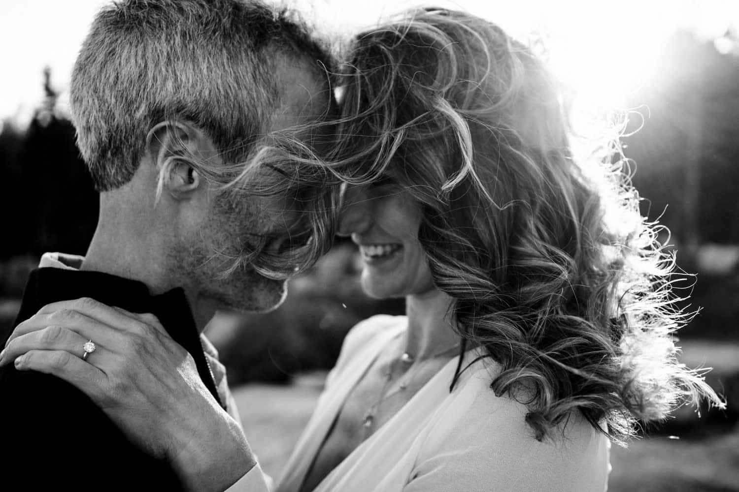 Black and white photo of a couple smiling and pressing their foreheads together, with the woman's curly hair blowing in the wind and the sun shining in the background.