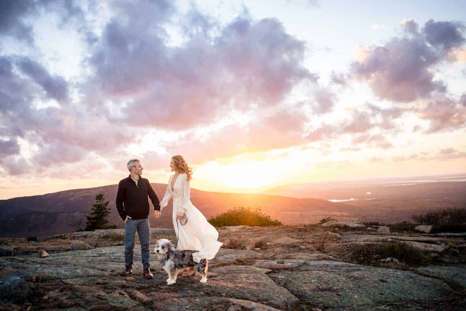 A couple holds hands and stands with a dog on a rocky landscape at sunset, with mountainous terrain and a dramatic sky in the background.