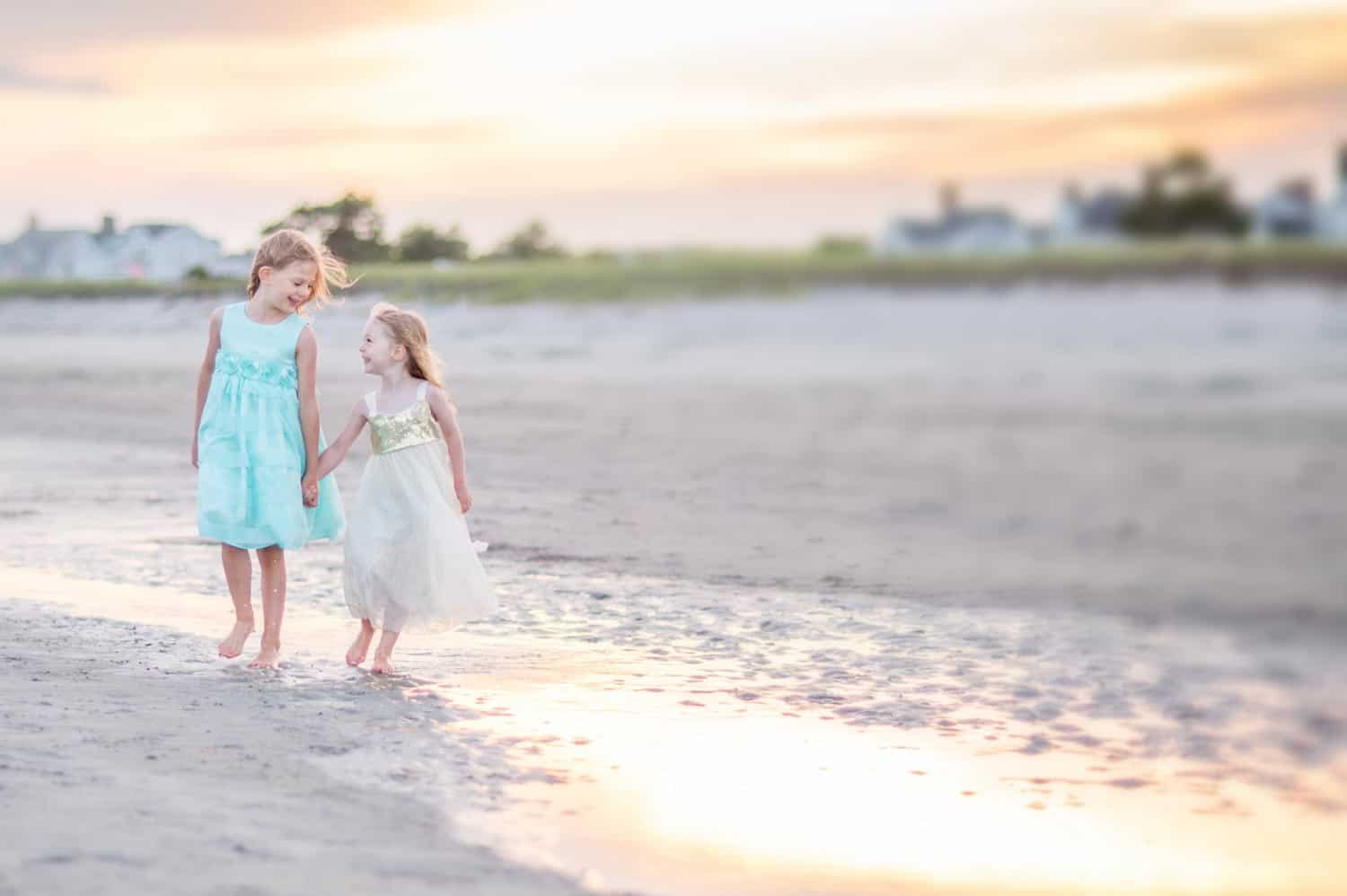 Two young girls in light-colored dresses walk hand-in-hand along a beach at sunset, with water and buildings in the background.