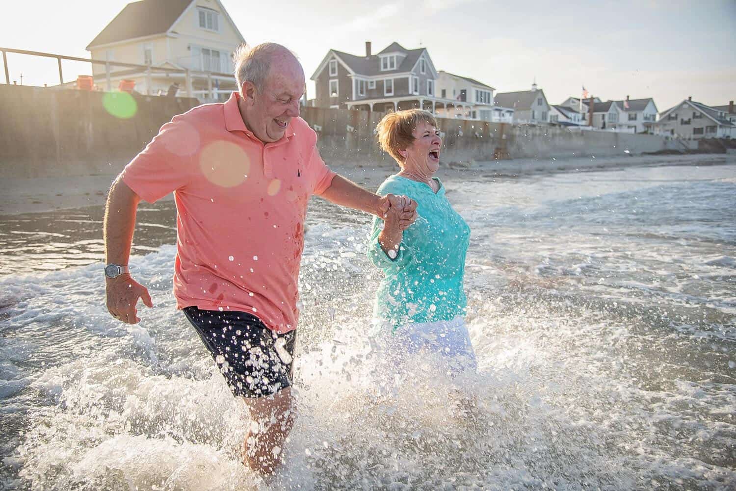 An elderly couple gleefully runs through the shallow water at a beach, holding hands. Houses and a stone retaining wall are visible in the background.