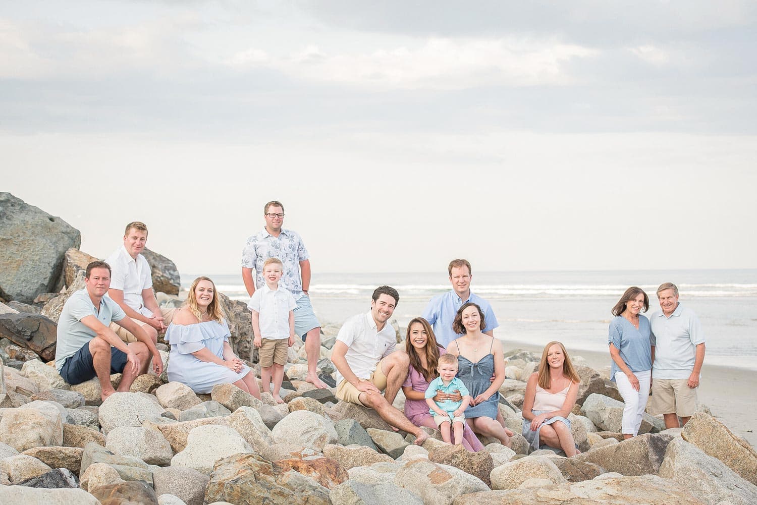 A group of people, including several children, sitting and standing on rocks by the beach. The sky is cloudy, and the ocean is in the background.