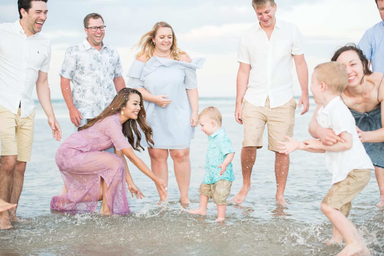 A group of adults and two children stand and play in shallow water at the beach, smiling and enjoying the moment.